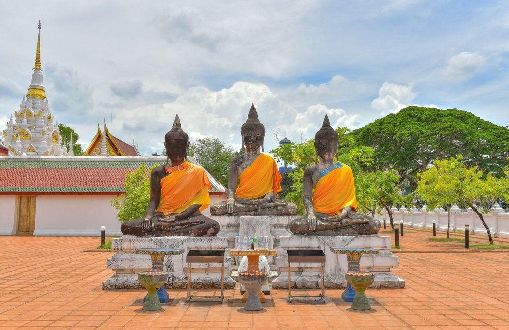 พระพุทธรูป 3 องค์คลุมผ้าสีส้ม เขื่อนเชี่ยวหลาน นั่งบนแท่นในลานวัด มีเจดีย์และต้นไม้ชวนให้นึกถึงภูมิทัศน์อันเงียบสงบใกล้กับเขื่อนรัชชประภาใต้ท้องฟ้าที่มีเมฆมาก