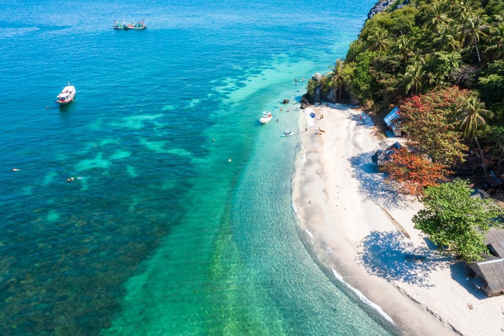 มุมมองทางอากาศของชายหาดเขตร้อนใน ชุมพรที่เที่ยว น้ำทะเลสีฟ้าใส เรือที่อยู่ไกลๆ ทรายสีขาว และต้นปาล์มสีเขียวชอุ่มเรียงรายอยู่ริมชายฝั่ง