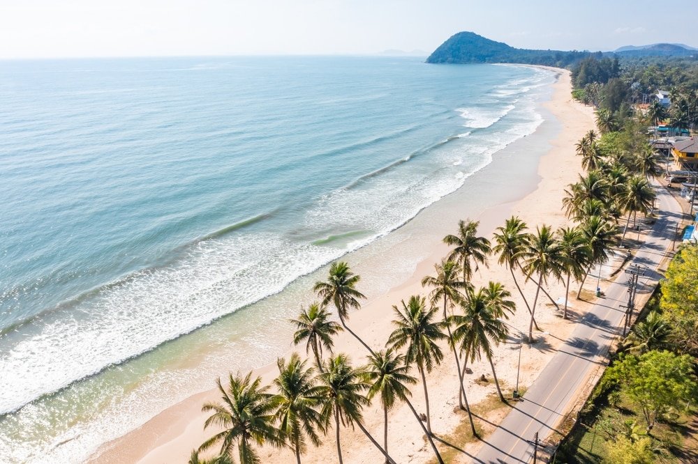 มุมมองทางอากาศของชายหาดเขตร้อนที่มีต้นปาล์ม คลื่นเบาๆ และเนินเขาไกลๆ ใต้ท้องฟ้าใสใน ชุมพรที่เที่ยว ถนนเลียบไปตามชายฝั่งที่งดงาม