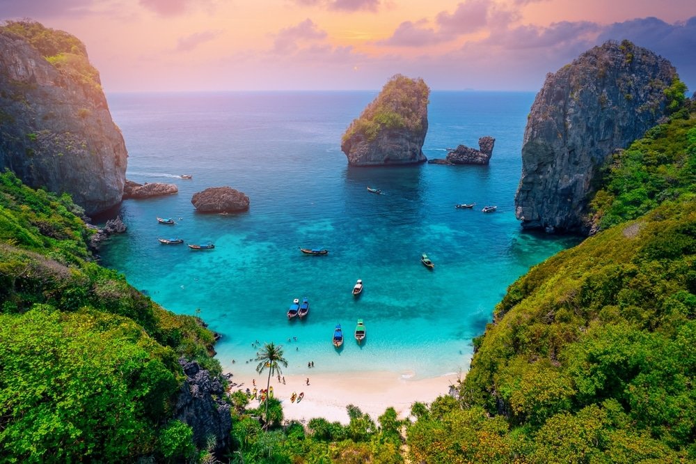 มุมมองทางอากาศ เกาะพีพี ของชายหาดเขตร้อนบนเกาะพีพี น้ำทะเลสีฟ้าใสล้อมรอบด้วยหน้าผาหินขนาดใหญ่และต้นไม้เขียวขจีภายใต้ท้องฟ้ายามพระอาทิตย์ตกที่มีสีสันสวยงาม เรือจอดทอดสมออยู่ใกล้ชายฝั่ง