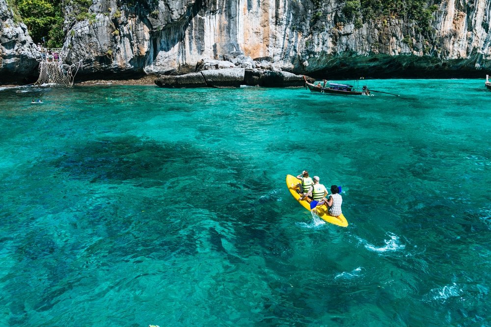 คนสองคนพายเรือคายัคสีเหลือง เกาะพีพี ในน้ำทะเลสีฟ้าใสใกล้หน้าผาหินอันน่าทึ่งของเกาะพีพี เรือและนักว่ายน้ำในระยะไกลช่วยเพิ่มความมีชีวิตชีวาให้กับทัศนียภาพบนเกาะที่สวยงามแห่งนี้ ดึงดูดทั้งผู้ที่แสวงหาการผจญภัยและผู้ที่รักธรรมชาติ