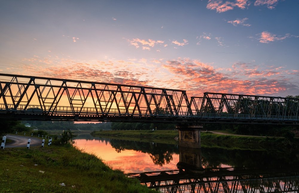 สะพานโครงเหล็กตั้งอยู่ใจกลางย่านเที่ยวชุมพร ทอดข้ามแม่น้ำอันเงียบสงบอย่างสง่างาม ที่เที่ยวชุมพร สะท้อนสีสันสดใสของท้องฟ้ายามพระอาทิตย์ตกดิน