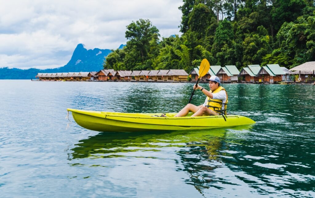 บุคคลในเรือคายัคสีเหลือง เขื่อนเชี่ยวหลาน ล่องลอยข้ามทะเลสาบอันเงียบสงบ ล้อมรอบด้วยทิวทัศน์อันงดงามของกระท่อมและต้นไม้ โดยมีฉากหลังอันน่าทึ่งของเขื่อนเชี่ยวหลาน