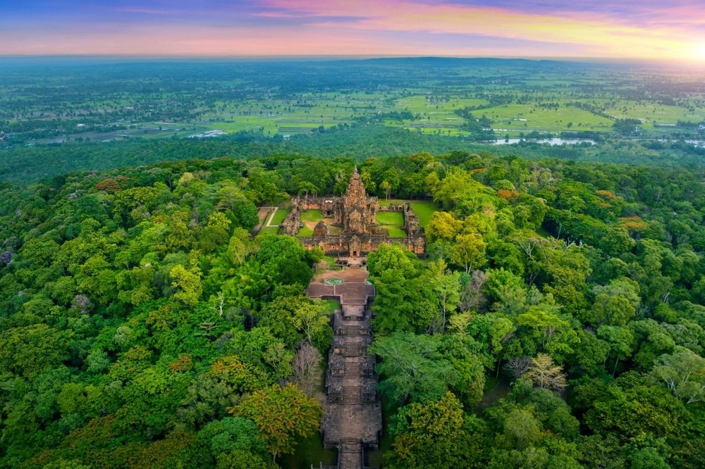 มุมมองทางอากาศของกลุ่มวัดประวัติศาสตร์บนเกาะพีพี ที่ล้อมรอบไปด้วยป่าเขียวขจีและทุ่งหญ้าใต้ท้องฟ้ายามพระอาทิตย์ตกที่มีสีสัน