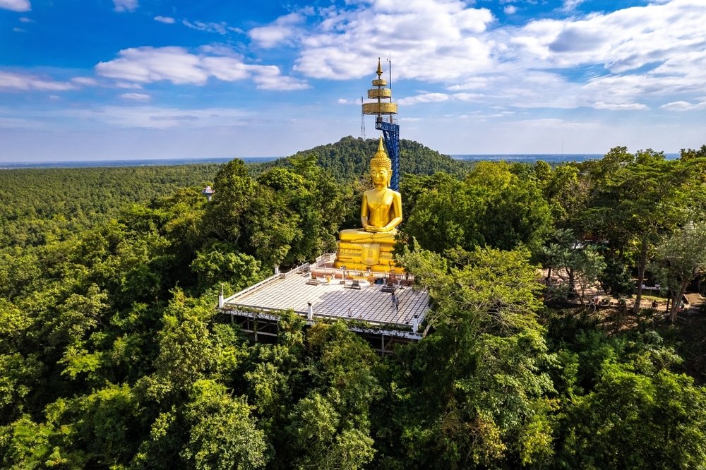 พระพุทธรูปทองคำองค์ใหญ่ประทับอยู่บนยอดเขาที่ปกคลุมด้วยป่าไม้ในเกาะพีพีจังหวัด โดยมีหอสื่อสารสูงอยู่ใกล้ๆ ใต้ท้องฟ้าที่มีเมฆบางส่วน