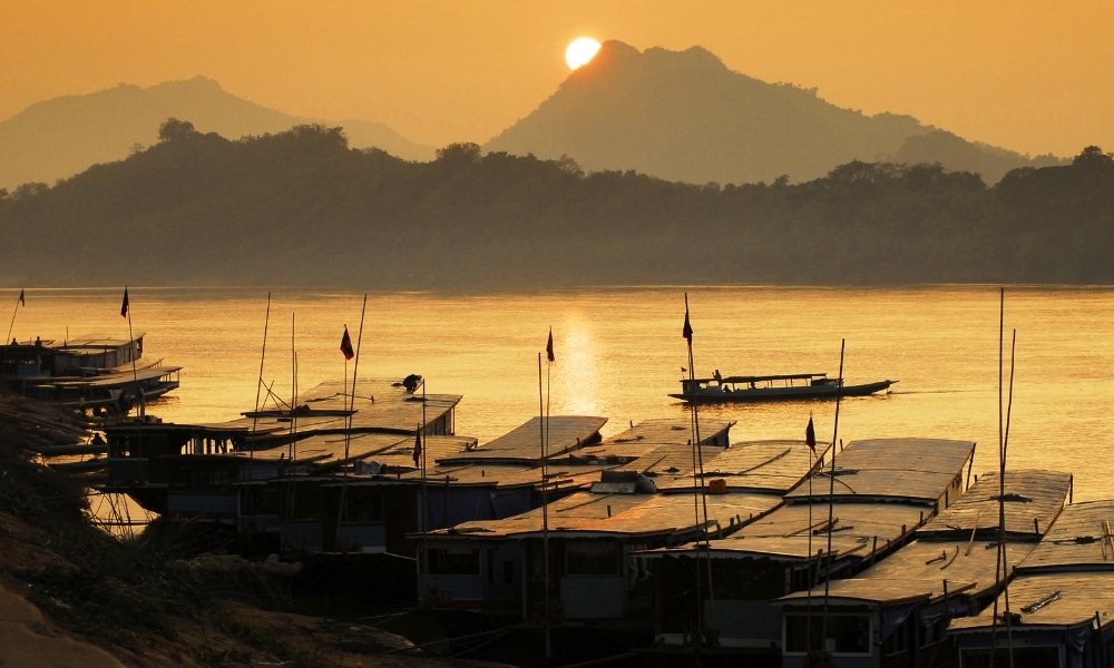 เรือที่เรียงรายไปตาม ท่องเที่ยวอีสาน ริมฝั่งแม่น้ำยามพระอาทิตย์ตก โดยมีภูเขาสูงตระหง่านเป็นฉากหลัง ทำให้เกิดฉากอันงดงามที่รวบรวมความเงียบสงบของท่องเที่ยวอีสาน.