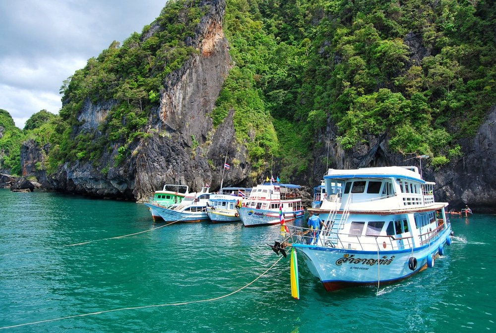 เรือจอดเทียบท่าอยู่ใกล้หน้าผาหินเขียวชอุ่มบนน้ำทะเลสีฟ้าครามใสภายใต้ ที่เที่ยวประเทศไทย ท้องฟ้าที่มีเมฆมาก มองเห็นความงามตามธรรมชาติของที่เที่ยวประเทศไทย