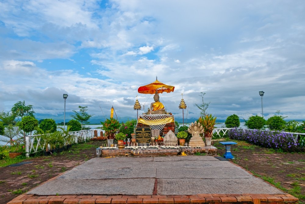 พระพุทธรูปสีทองอร่ามอยู่ใต้ร่มสีแดงและเหลือง ล้อมรอบด้วยเครื่องเซ่น  ที่เที่ยวประเทศไทย ประดิษฐานอยู่บนแท่นประดับตกแต่งในหนึ่งในจุดที่ต้องมาเยี่ยมชมเมื่อมา เที่ยวไทย โดยมีฉากหลังเป็นต้นไม้เขียวขจีและท้องฟ้าที่มีเมฆปกคลุม