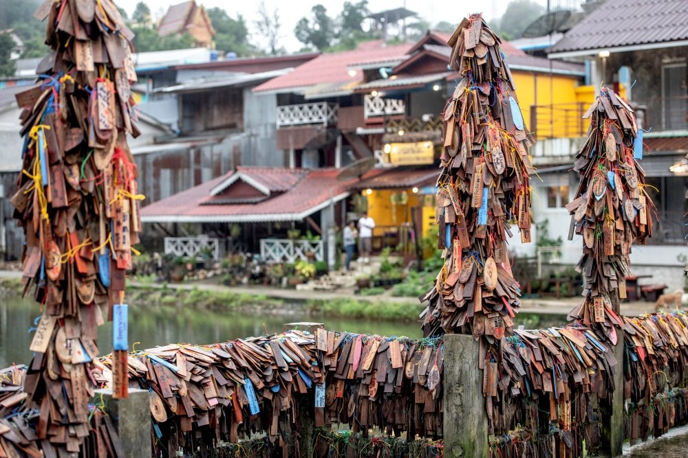 ป้ายไม้ประดับแขวนตามเสาและรั้วในหมู่บ้าน โดยมีบ้านเรือนและต้นไม้เขียวขจีเป็นฉากหลัง สะท้อนถึงเสน่ห์ของที่เที่ยวประเทศไทย เที่ยวไทย 