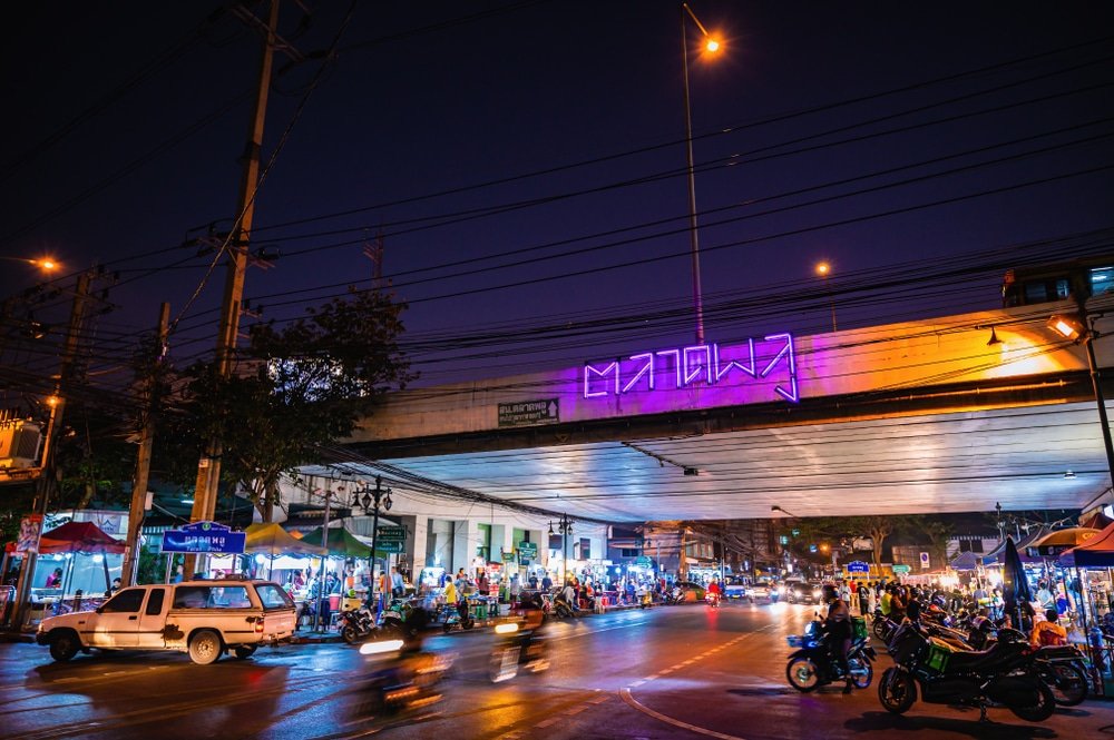 ตลาดกลางคืนอันพลุกพล่านเกิดขึ้นภายใต้สะพานลอยทางหลวงที่ติดไฟในที่เที่ยวประเทศไทย พร้อมด้วย เที่ยวไทย ยานพาหนะที่กำลังเคลื่อนที่ แผงลอยที่ส่องสว่าง และผู้คนที่เดินไปตามถนนที่เปียกชื้น ภาพถ่ายที่มีชีวิตชีวานี้รวบรวมเสน่ห์อันน่าหลงใหลของเที่ยวไทย