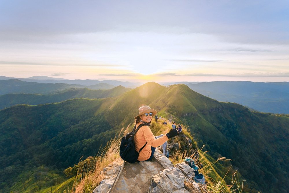 คนกำลังนั่งอยู่บนหิน ที่เที่ยวประเทศไทย ยื่นออกไปบนขอบเขา สวมหมวกและถือกล้องถ่ายรูป พระอาทิตย์กำลังตกเหนือเนินเขาสีเขียวขจี จับภาพทิวทัศน์ที่สวยงามตระการตาที่มักดึงดูดผู้ที่รักการท่องเที่ยวไทย