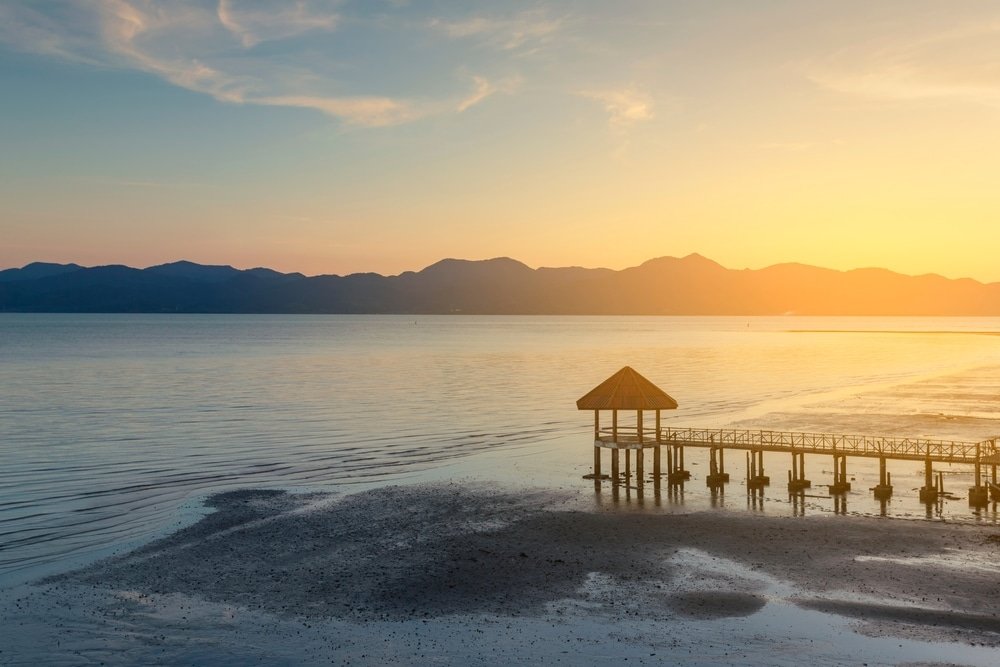 ท่าเรืออันเงียบสงบพร้อมที่พักเล็กๆ ที่เที่ยวประเทศไทย  ทอดยาวลงไปในผืนน้ำอันสงบยามพระอาทิตย์ตก มีภูเขาอยู่ไกลๆ และชายหาดทรายอยู่เบื้องหน้า นับเป็นภาพที่สมบูรณ์แบบของเที่ยวไทย