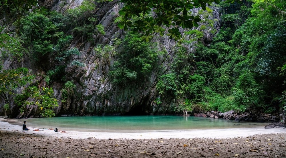 ชายหาดอันเงียบสงบที่มีหาดทรายขาวและน้ำทะเลใส ล้อมรอบด้วย ที่เที่ยวประเทศไทย หน้าผาหินและใบไม้สีเขียวหนาแน่น เป็นจุดที่สมบูรณ์แบบสำหรับผู้ที่ต้องการเที่ยวไทย สองคนกำลังพักผ่อนบนผืนทราย เพลิดเพลินกับอัญมณีที่ซ่อนอยู่ในหมู่นักท่องเที่ยวที่เที่ยวประเทศไทย