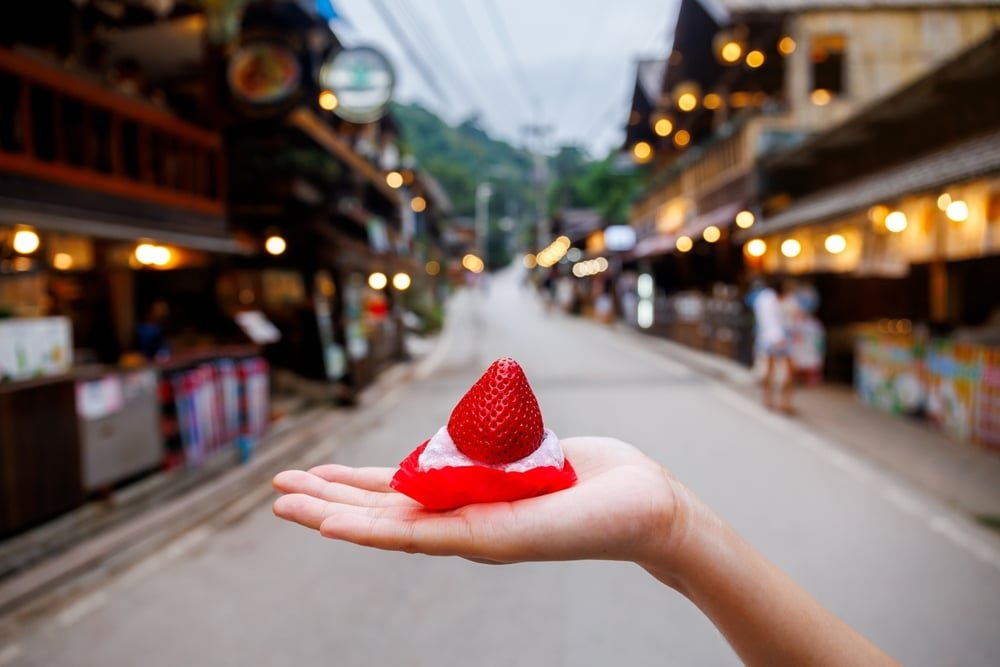 มือถือขนมหวานชิ้นเล็กๆ ที่ราดด้วยสตรอเบอร์รี่ไว้หน้าถนนที่มีร้านค้าเรียงราย สะท้อนถึงเสน่ห์ของเที่ยวไทย เที่ยวไทย 