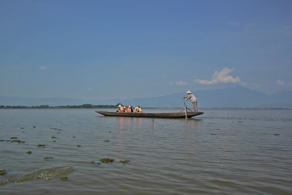 ชายคนหนึ่งกำลังพายเรือยาวใน ที่เที่ยวประเทศไทย ทะเลสาบอันเงียบสงบภายใต้ท้องฟ้าสีฟ้าใส โดยมีภูเขาอันไกลโพ้นอยู่เบื้องหลัง เป็นภาพที่ทำให้หวนนึกถึงการท่องเที่ยวประเทศไทย ผู้โดยสารสามคนนั่งอยู่ที่หัวเรือ ท่ามกลางความเงียบสงบของธรรมชาติ