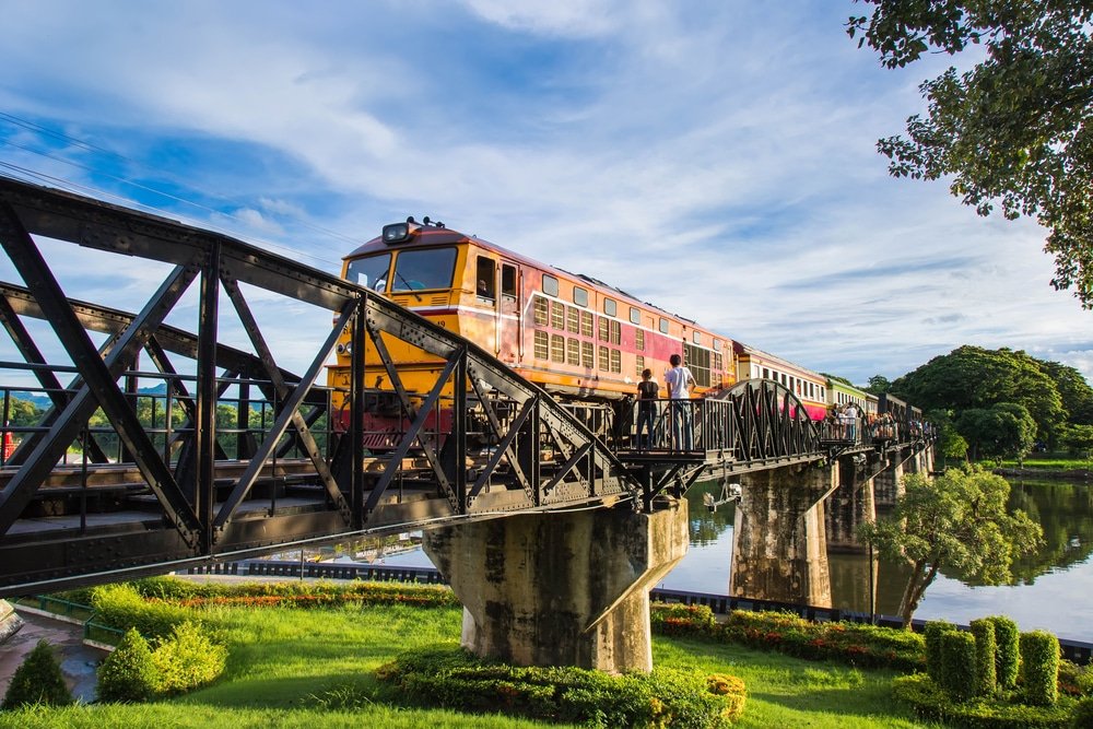 รถไฟวิ่งข้ามสะพานเหล็กที่รายล้อมไปด้วยต้นไม้เขียวขจีและน้ำระยิบระยับใต้ท้องฟ้าสีฟ้า ถ่ายทอดความ เที่ยวไทย งามอันน่าหลงใหลของประเทศไทยได้เป็นอย่างดี นับเป็นภาพที่สมบูรณ์แบบของการท่องเที่ยวไทยที่ธรรมชาติและความทันสมัยผสมผสานกันอย่างลงตัว