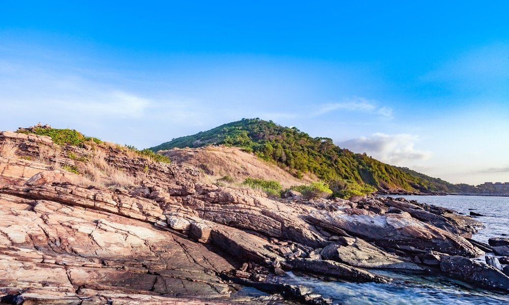 ชายหาดหินมีเนินหญ้าริมน้ำภายใต้ ที่เที่ยวระยอง ท้องฟ้าสีฟ้าใส เหมาะกับผู้แสวงหาความงามทางธรรมชาติของระยองที่เที่ยว.