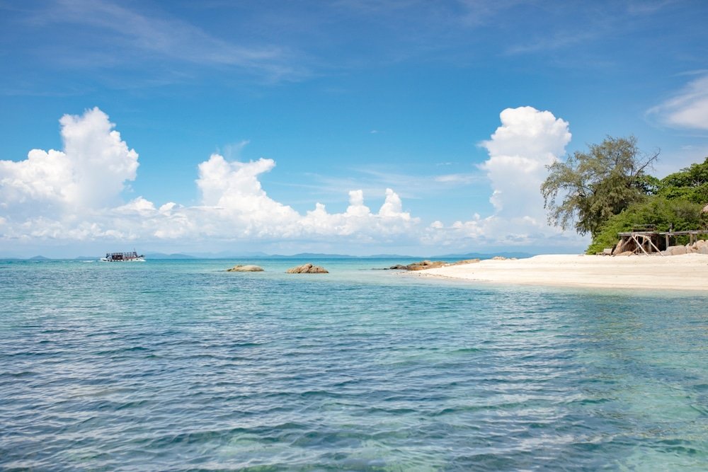 ชายหาดอันเงียบสงบในที่เที่ยวระยองมีน้ำทะเลสีฟ้าใส หาดทราย และต้นไม้เล็กๆ สองสามต้นใต้ท้องฟ้าสดใสและมีเมฆบางส่วน มองเห็นเรือลำ ที่เที่ยวระยอง หนึ่งอยู่บนขอบฟ้า สะท้อนถึงแก่นแท้ของสถานที่ท่องเที่ยวอันเงียบสงบแห่งนี้.