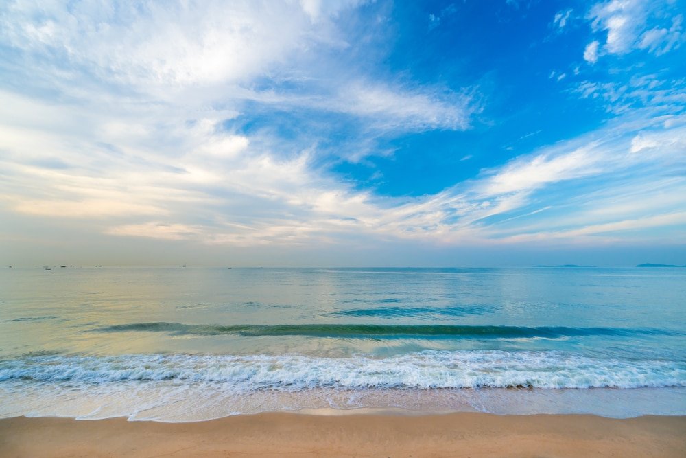 สัมผัสบรรยากาศชายหาดอันเงียบสงบในระยองที่เที่ยว ซึ่งมีคลื่นซัดสาด สถานที่เที่ยวระยอง ชายฝั่งภายใต้ท้องฟ้าสีครามสดใสที่ประดับประดาด้วยเมฆที่โปรยปราย.