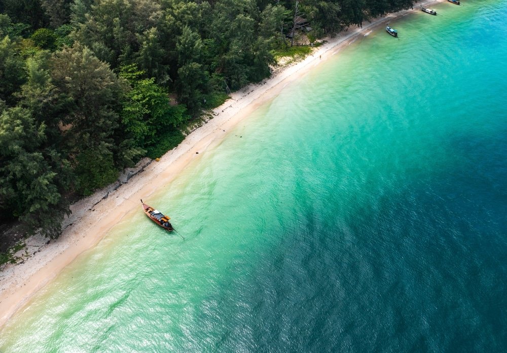 มุมมองทางอากาศของเรือเล็กใกล้หาดทรายที่ล้อมรอบด้วยต้นไม้สีเขียวชอุ่ม ท่องเที่ยวตรัง และน้ำทะเลสีฟ้าครามใส จับภาพความงามอันเงียบสงบชวนให้นึกถึงท่องเที่ยวอีสาน.