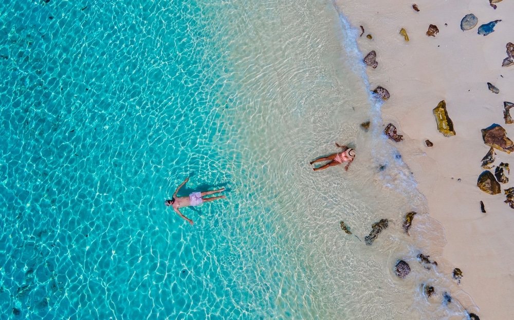 คนสองคนผ่อนคลายที่ชายหาดอัน ท่องเที่ยวตรัง เงียบสงบ คนหนึ่งลอยอยู่ในน้ำทะเลสีฟ้าครามใส ในขณะที่อีกคนพักผ่อนบนหาดทรายท่ามกลางโขดหินที่กระจัดกระจาย ชวนให้นึกถึงจุดหมายปลายทางที่พบในพื้นที่ท่องเที่ยวภาคอีสาน10อันดับ.