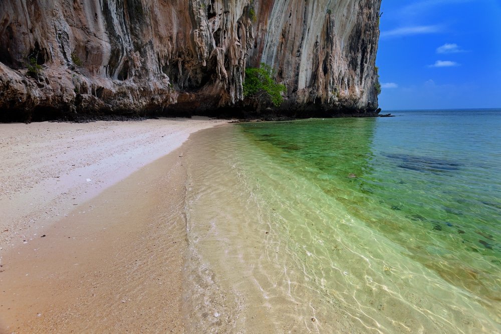 หาดทรายที่มีน้ำตื้นชัดเจนข้าง ท่องเที่ยวตรัง หน้าผาหินปูนสูงตระหง่านใต้ท้องฟ้าสีครามเป็นสถานที่พักผ่อนที่งดงาม อัญมณีที่ซ่อนอยู่นี้เป็นหนึ่งในแหล่งท่องเที่ยวในอีสานที่ดึงดูดนักท่องเที่ยวที่แสวงหาความเงียบสงบและความงามตามธรรมชาติ.