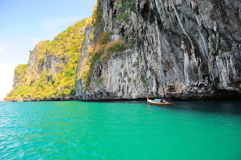 เรือหางยาวแล่นผ่านน้ำทะเลสีฟ้าครามใสใกล้หน้าผาหินขนาดใหญ่ที่ประดับประดาด้วยพืชพรรณสีเขียว ท่องเที่ยวตรัง ชอุ่มชวนให้นึกถึงความงามอันเงียบสงบที่พบในแหล่งท่องเที่ยวในอีสาน.