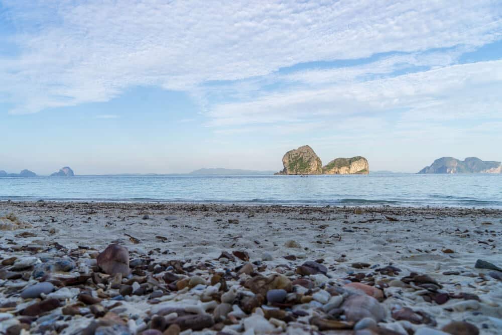 หาดทรายอันเงียบสงบที่มีก้อนกรวดกระจัดกระจายอยู่เบื้องหน้า ทะเลอัน ท่องเที่ยวตรัง เงียบสงบทอดยาวไปไกล และเกาะหินภายใต้ท้องฟ้าที่มีเมฆมาก ชวนให้นึกถึงความงามอันเงียบสงบที่พบในจุดหมายปลายทางอันเงียบสงบในอุทยานท่องเที่ยวภาคอีสาน.