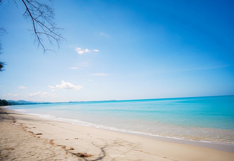 ฉากชายหาดอันเงียบสงบในเขาหลักที่เที่ยวกับท้องฟ้าสีฟ้าใส ท้องทะเลสีฟ้าคราม และหาดทรายที่ว่างเปล่า กิ่งก้านของต้นไม้กระจัดกระจายอยู่ทางด้านซ้าย เป็นจุดที่เหมาะสำหรับการสะท้อนและผ่อนคลาย.