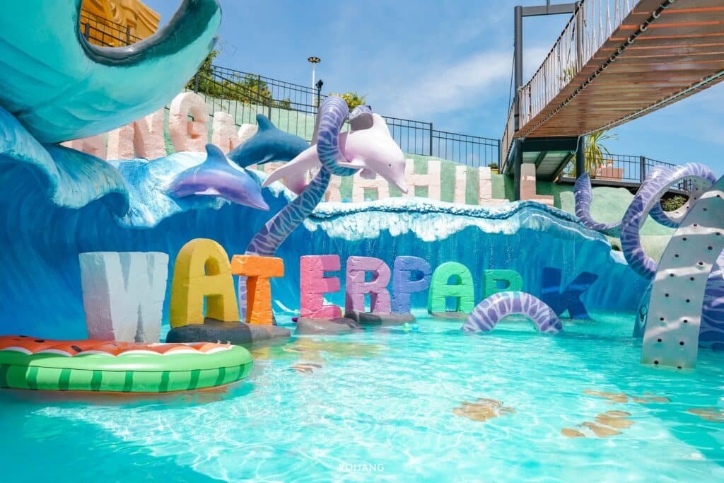 ทางเข้าสวนน้ำที่มีชีวิตชีวา ประดับด้วยตัวอักษร "WATERPARK" ตัวหนา ล้อมรอบด้วยคลื่นสีฟ้าร่าเริง โลมาขี้เล่น หนวดที่หมุนวน และห่วงยางลอยน้ำ ทั้งหมดนี้อยู่ใต้ท้องฟ้าสดใสใกล้กับทะเล.