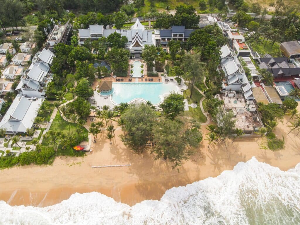 มุมมองทางอากาศของรีสอร์ทริมชายหาดที่มีลักษณะ เสม็ดนางชีที่พัก มีสระว่ายน้ำกลางขนาดใหญ่ที่ล้อมรอบด้วยต้นไม้เขียวขจีและอาคารทันสมัย พร้อมด้วยคลื่นซัดชายฝั่งทราย.