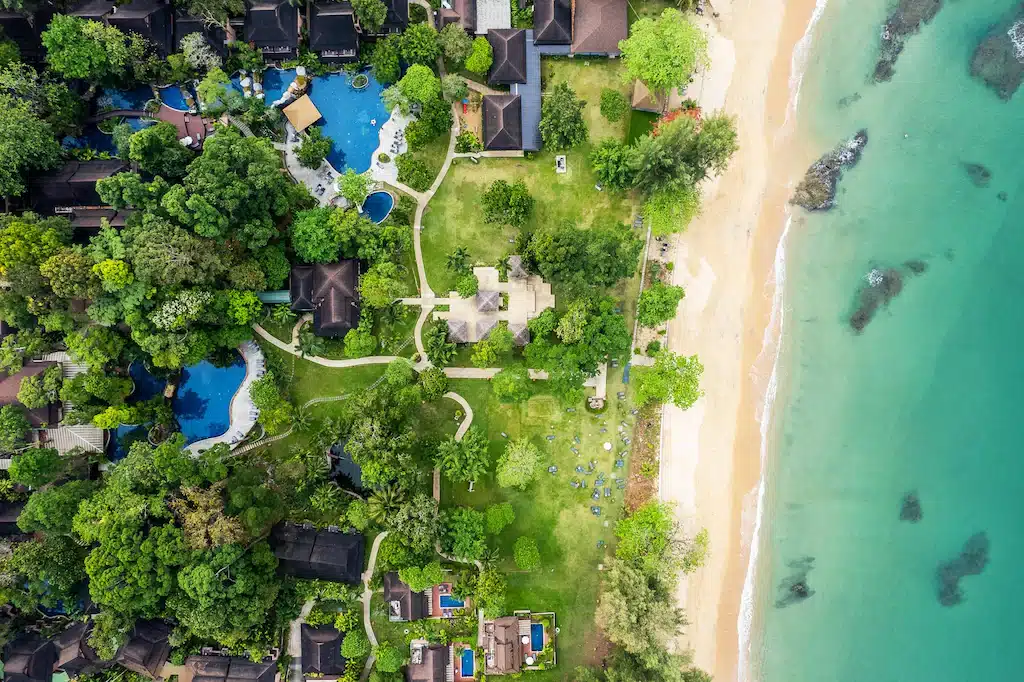 มุมมองทางอากาศของรีสอร์ทริมชายหาด ฟังก์ชั่นพังงาติดทะเล พร้อมสวนเขียวชอุ่ม สระว่ายน้ำที่น่าดึงดูดใจ และทางเดินคดเคี้ยวที่นำไปสู่หาดทรายและมหาสมุทรสีฟ้าครามที่ส่องประกายระยิบระยับ.