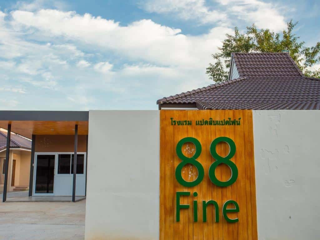 อาคารที่มีป้าย "88 Fine" บนแผงไม้ตั้งอยู่ติดกับผนังสีขาวเตี้ยๆ มุมมองชั้นดาดฟ้าและบางส่วนของโครงสร้างสมัยใหม่ที่ทอดยาวภายใต้ท้องฟ้าสีฟ้าใส ผสมผสานเสน่ห์ของ โรงแรมสุราษฎร์ธานี.