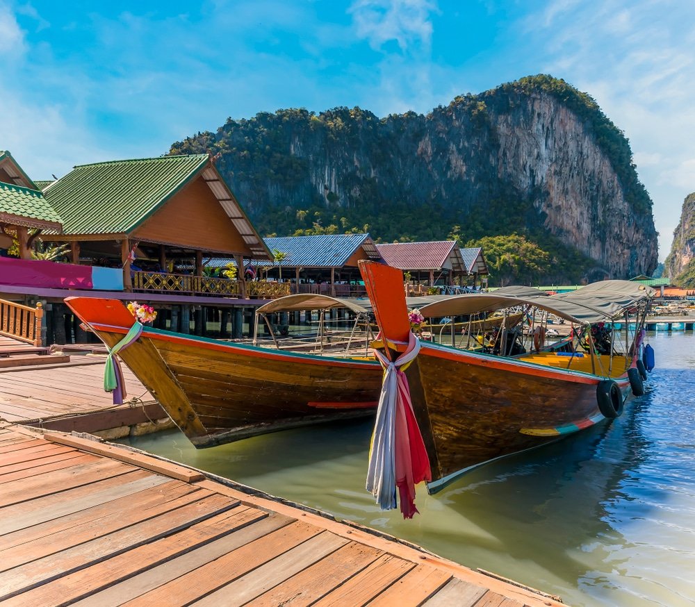 เรือหางยาวสองลำที่ปูด้วยผ้าสีสันสดใสจอดอยู่ที่ท่าเรือไม้บน เกาะปันหยี ซึ่งล้อมรอบด้วยบ้านใต้ถุนบ้าน และมีเนินหินขนาดใหญ่เป็นฉากหลังภายใต้ท้องฟ้าสีฟ้าใส.