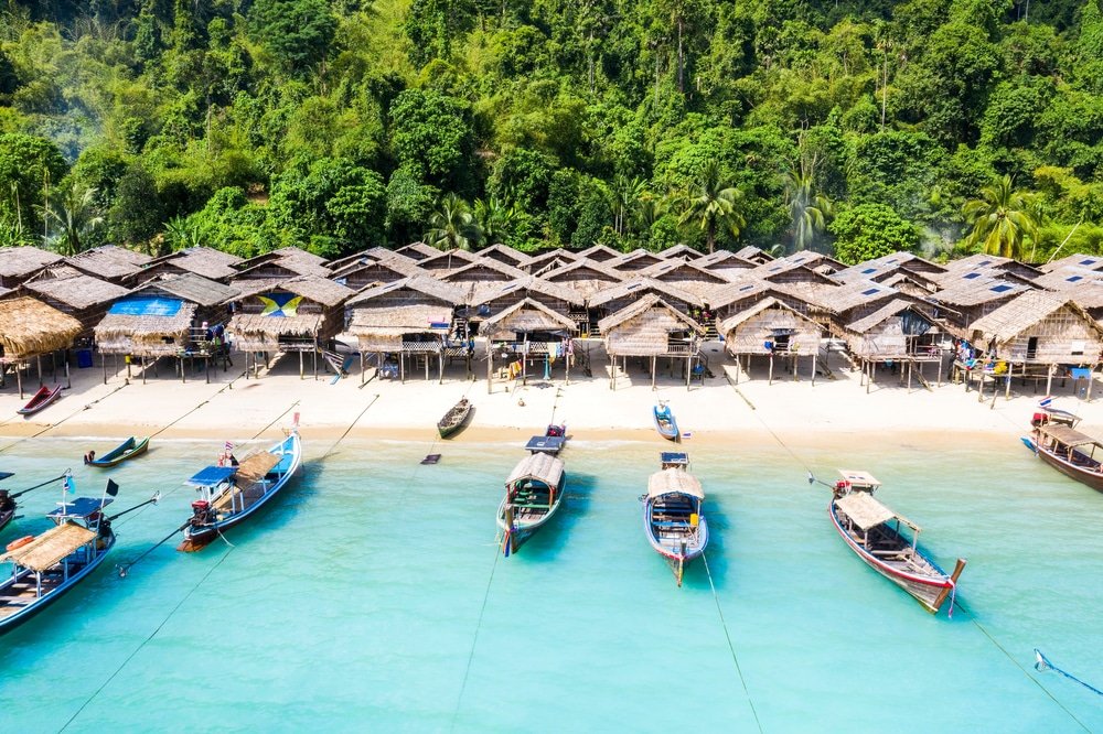 มุมมองทางอากาศของกระท่อม เกาะปันหยี หลังคาฟางที่เรียงรายอยู่ริมชายหาดบนเกาะปันหยี พร้อมด้วยเรือสีสันสดใสหลายลำที่จอดทอดสมออยู่ในน้ำทะเลสีฟ้าใสและมีป่าสีเขียวขจีเป็นฉากหลัง.