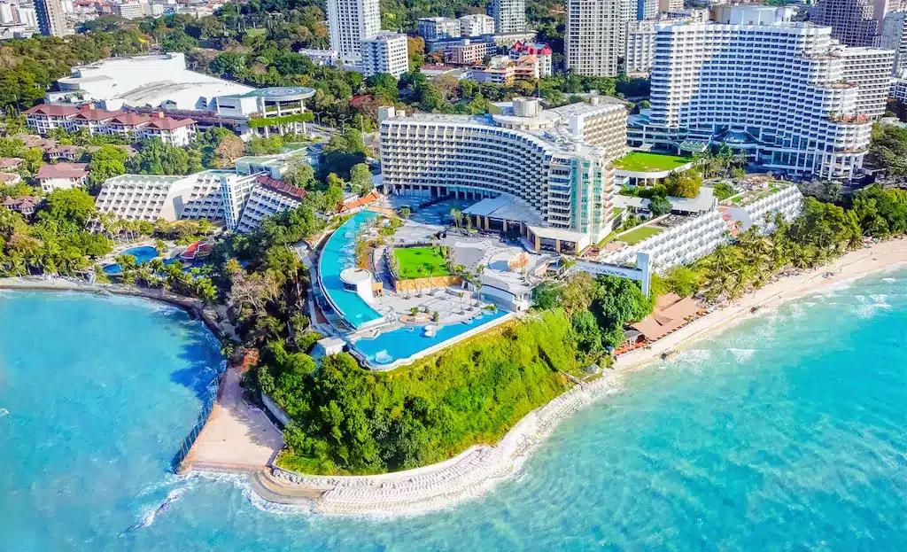รีสอร์ทริมชายหาดขนาดใหญ่ที่มีอาคารหลายหลัง สระว่ายน้ำ และสวน ตั้งอยู่บนคาบสมุทรสีเขียวติดกับน้ำทะเลสีฟ้าใส เหมาะสำหรับผู้แสวงหาความผ่อนคลายและความหรูหราริมทะเล พัทยาที่เที่ยว. ที่เที่ยวพัทยา 
