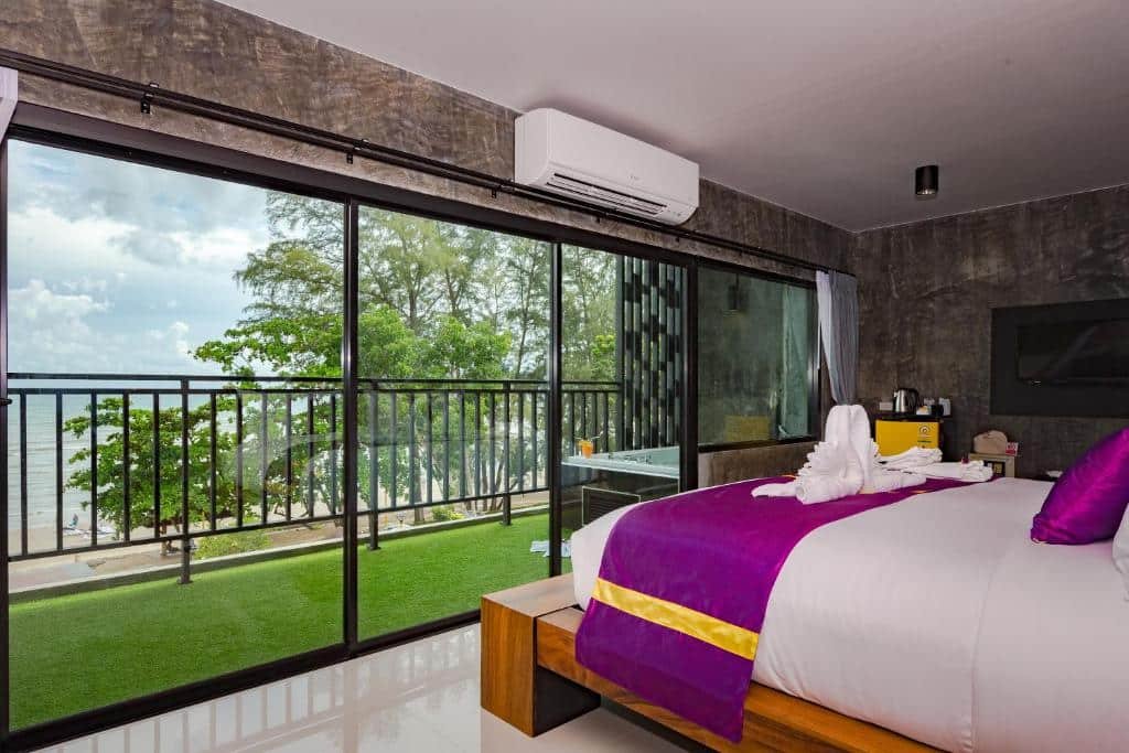 ห้องพักโรงแรมทันสมัยที่ รีสอร์ทกระบี่  มีประตูกระจกบานใหญ่เปิดออกสู่ระเบียงมองเห็นต้นไม้และทะเล เตียงนอนมีเครื่องนอนสีขาวและสีม่วงพร้อมภาพศิลปะผ้าเช็ดตัวอยู่ด้านบน.