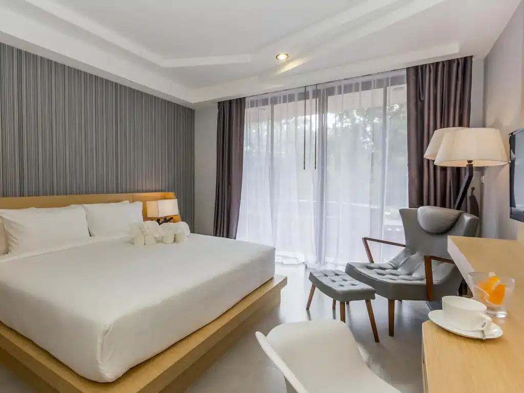 ห้องพักในโรงแรม ที่พักกระบี่ ทันสมัยพร้อมเตียงขนาดใหญ่ ผนังเน้นสีเทา เก้าอี้เท้าแขนแสนสบายพร้อมออตโตมัน โต๊ะทำงาน และหน้าต่างสูงจากพื้นจรดเพดาน เหมาะสำหรับผู้ที่กำลังมองหาที่พักสไตล์กระบี่ที่มีสไตล์หรือสัมผัสประสบการณ์รีสอร์ทกระบี่ที่สะดวกสบาย.