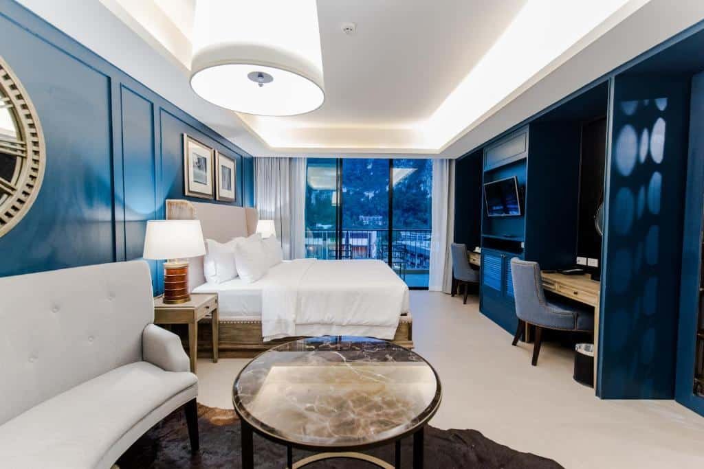 ห้องพักโรงแรมทันสมัยในโรงแรมเมืองกระบี่ที่มีสไตล์ พร้อมผนังสีฟ้า เตียงนอนนุ่มสบาย ผ้าปูที่นอนสีขาว พื้นที่นั่งเล่นแสนสบาย โต๊ะหินอ่อน  โรงแรมกระบี่ โต๊ะทำงาน ทีวีติดผนัง หน้าต่างบานใหญ่ และระเบียงที่มองเห็นทิวทัศน์ภูเขาอันสวยงาม.