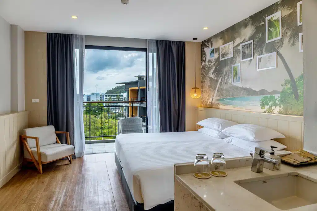 ห้องพักในโรงแรม ที่พักกระบี่ ทันสมัยพร้อมเตียงคู่ ระเบียง เก้าอี้ และภาพจิตรกรรมฝาผนังธีมเมืองร้อน อ่างล้างหน้าและแก้วน้ำที่มองเห็นได้ในเบื้องหน้า เหมาะสำหรับการพักผ่อนอย่างผ่อนคลายในโรงแรมกระบี่.