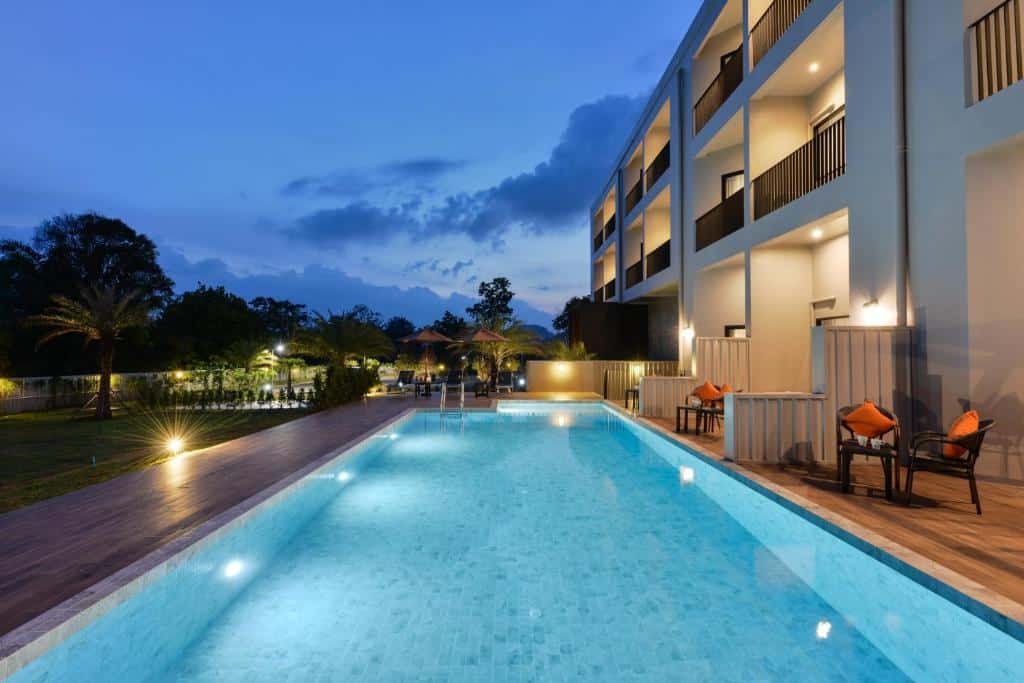 รีสอร์ทกระบี่ โรงแรมกระบี่ อันทันสมัยพร้อมระเบียงมองเห็นสระว่ายน้ำกลางแจ้งขนาดใหญ่ที่มีแสงสว่างส่องสว่างในเวลาพลบค่ำ ล้อมรอบด้วยเก้าอี้นั่งเล่นและภูมิทัศน์สวนอันเขียวชอุ่ม.