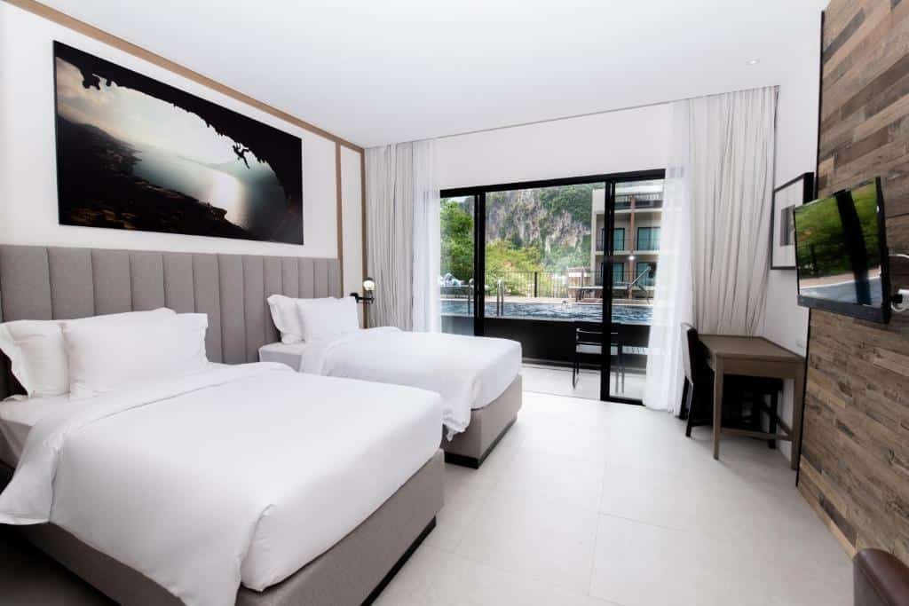 ห้องพักในโรงแรมทันสมัยพร้อมเตียงแฝดสองเตียง งานศิลปะบนผนังขนาดใหญ่ โต๊ะพร้อมเก้าอี้ ทีวี และประตูกระจกบาน โรงแรมกระบี่ เลื่อนที่เปิดออกสู่ระเบียงพร้อมวิวสระว่ายน้ำและกลางแจ้ง เหมาะสำหรับการเข้าพักที่โรงแรมกระบี่หรือรีสอร์ทกระบี่.