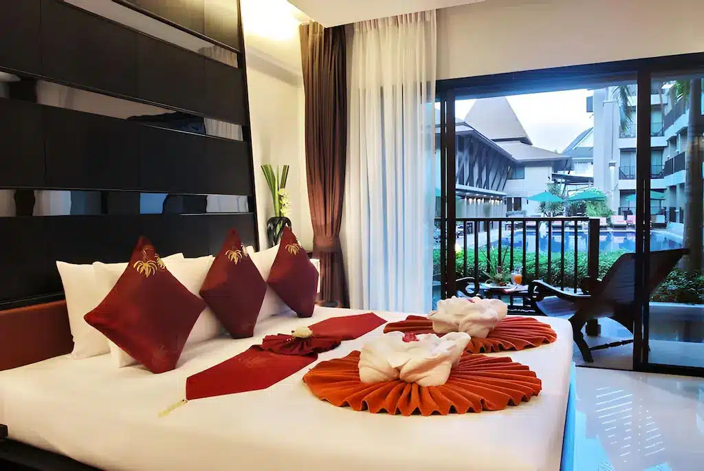 ห้องพัก ที่พักกระบี่ ในโรงแรมทันสมัยพร้อมเตียงขนาดใหญ่ หมอนอิงสีแดง และผ้าขนหนูพับเป็นรูปหงส์ ถัดจากประตูกระจกที่เปิดออกไปสู่ลานริมสระว่ายน้ำ เหมาะสำหรับการเข้าพักในรีสอร์ทสุดเก๋แห่งนี้.