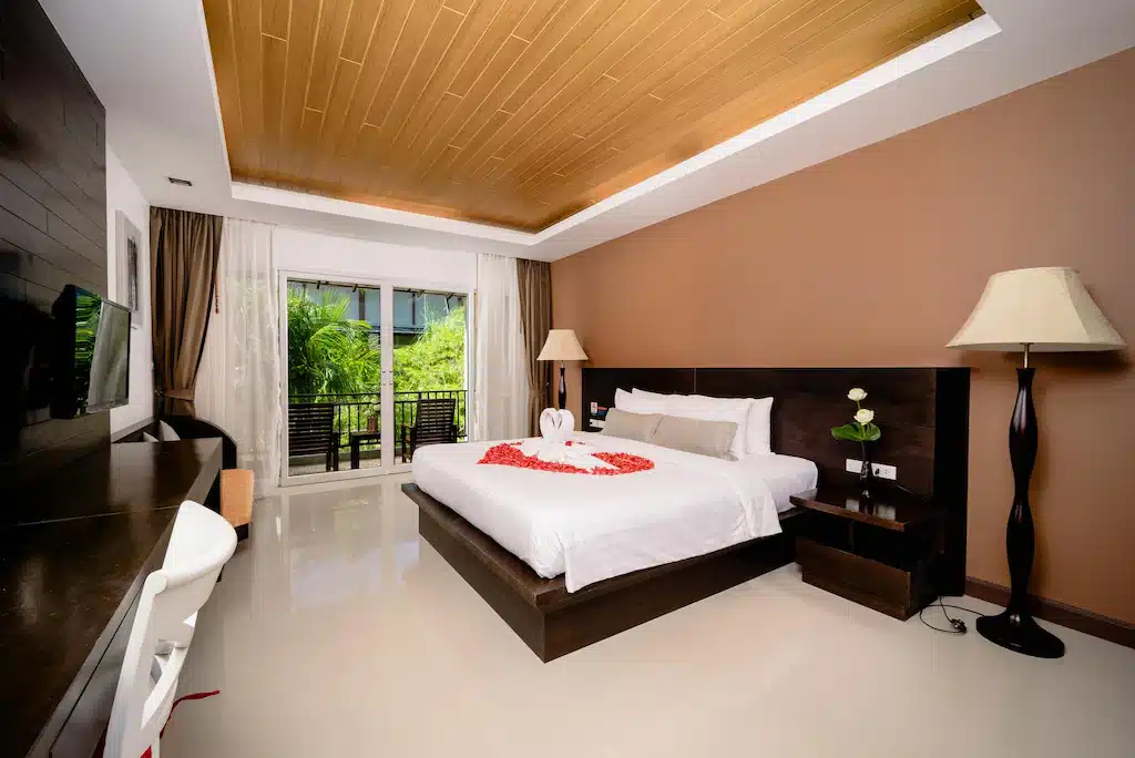 ห้องพักโรงแรมทันสมัยพร้อมเตียงขนาดใหญ่ตกแต่งด้วยผ้าขนหนูลาย ที่พักกระบี่ หงส์และกลีบกุหลาบ โต๊ะทำงาน ทีวี โคมไฟตั้งพื้น และระเบียงที่มองเห็นทิวทัศน์อันเขียวขจี เหมาะสำหรับการเข้าพักของคุณที่รีสอร์ทกระบี่.