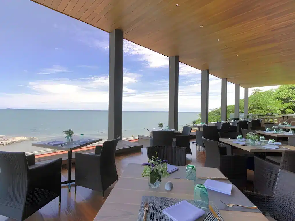 ระเบียงร้านอาหารแบบเปิดโล่งพร้อมโต๊ะและเก้าอี้ทันสมัยที่มองเห็นวิวทะเล มีเครื่องแก้วและผ้าเช็ดปากที่ ที่เที่ยวพัทยา จัดอย่างประณีตใต้เพดานไม้ เหมาะสำหรับเที่ยวพัทยาหรือเฉพาะที่เที่ยวพัทยาติดทะเล.