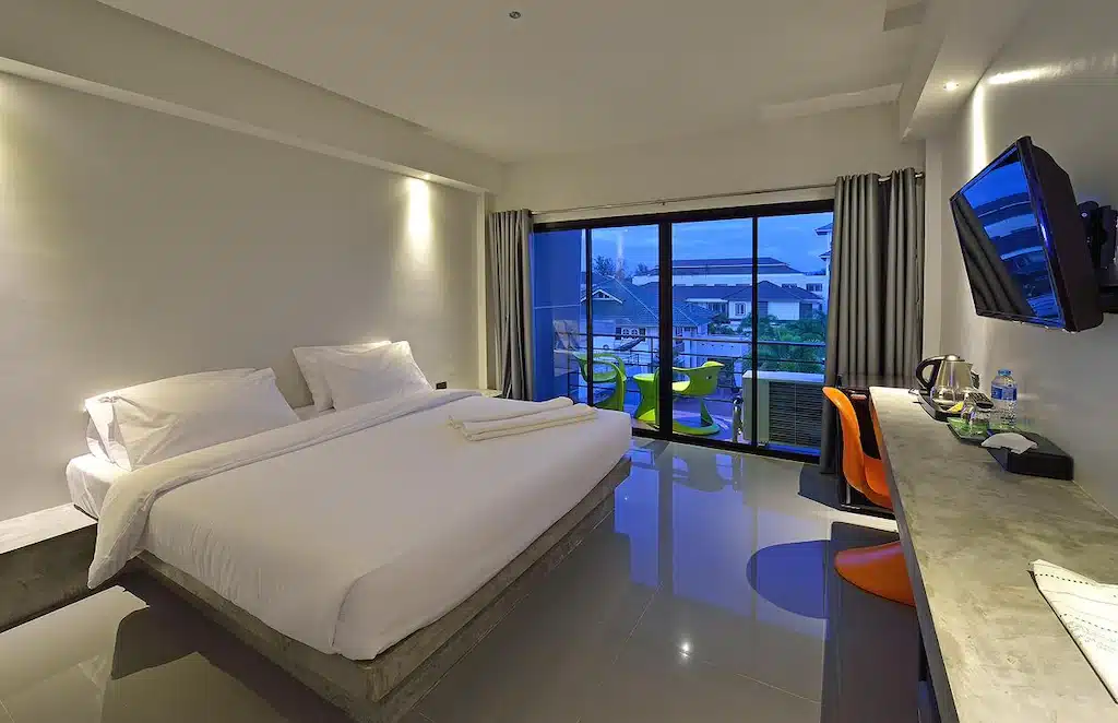 ห้องพักในโรงแรมทันสมัยพร้อมเตียง ที่พักกระบี่ คู่ ทีวีติดผนัง โต๊ะทำงาน และระเบียงพร้อมที่นั่งกลางแจ้งที่มองเห็นอาคารใกล้เคียง เหมาะสำหรับผู้ที่กำลังมองหาที่พักในเมืองกระบี่ที่มีสไตล์ในระหว่างการเดินทาง.