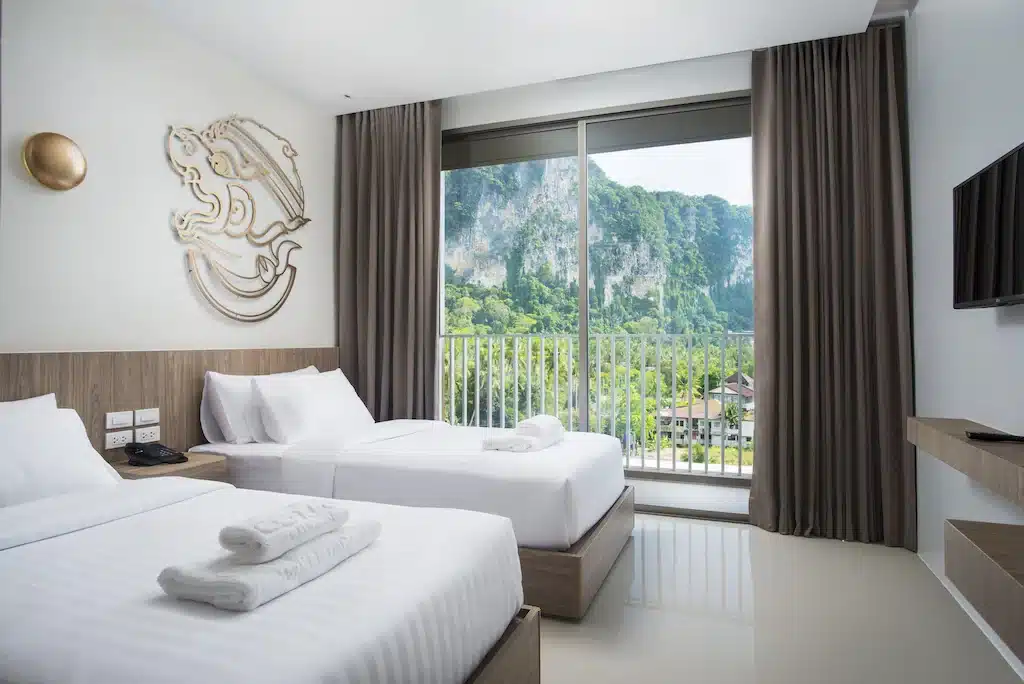 ห้องพัก ที่พักกระบี่ ในโรงแรมทันสมัยพร้อมเตียงเดี่ยว 2 เตียง ชุดเครื่องนอนสีขาว งานศิลปะบนผนัง ทีวีจอแบน และระเบียงพร้อมวิวภูเขาอันเขียวชอุ่ม เหมาะสำหรับผู้ที่กำลังมองหาที่พักรีสอร์ทกระบี่หรือกระบี่ในบรรยากาศธรรมชาติอันเงียบสงบ.