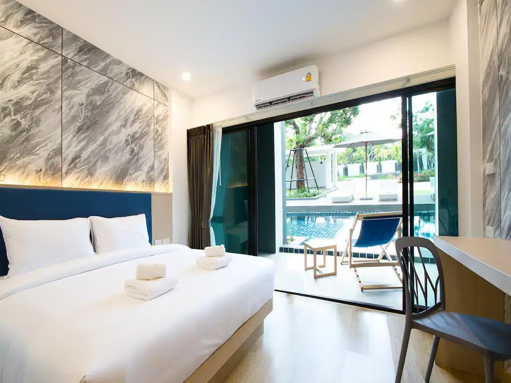 ห้องพักโรงแรมทันสมัยที่ รีสอร์ทกระบี่  มีเตียงขนาดใหญ่ โต๊ะทำงาน และประตูบานเลื่อนกระจกเปิดออกสู่ลานเฉลียงพร้อมวิวสระว่ายน้ำ ผ้าขนหนูพับไว้บนเตียง และแสงธรรมชาติส่องเข้ามาเติมเต็มพื้นที่อันน่าดึงดูดใจนี้.