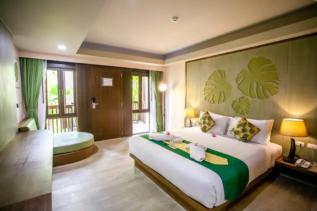 ห้องพักโรงแรมทันสมัยที่ โรงแรมกระบี่ พร้อมเตียงคู่ ตกแต่งด้วยสีเขียว โคมไฟข้างเตียง 2 โคมไฟ หน้าต่างบานใหญ่พร้อมผ้าม่าน และโต๊ะไม้ ผ้าเช็ดตัวที่พับไว้บนเตียงและการตกแต่งผนังอันร่มรื่นช่วยเพิ่มประสบการณ์การพักผ่อนที่กระบี่ของคุณ.