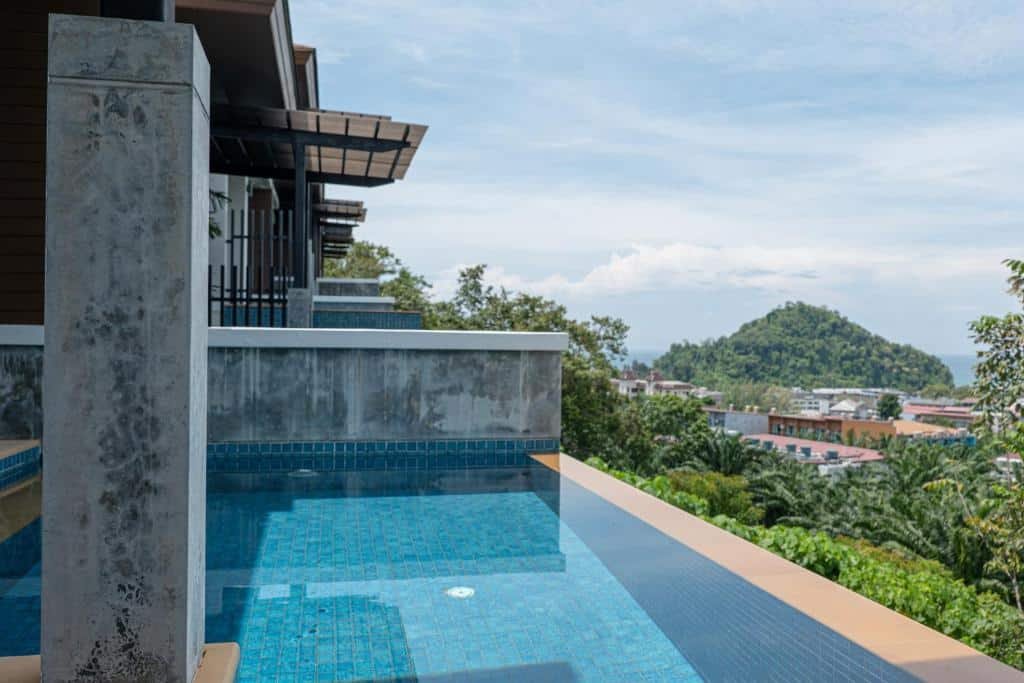 สระว่ายน้ำไร้ขอบบนระเบียงที่อ่าวนางกระบี่ พร้อมทิวทัศน์เนินเขา ต้นไม้อัน ที่พักอ่าวนาง เขียวชอุ่ม และอาคารต่างๆ ภายใต้ท้องฟ้าที่มีเมฆบางส่วน.